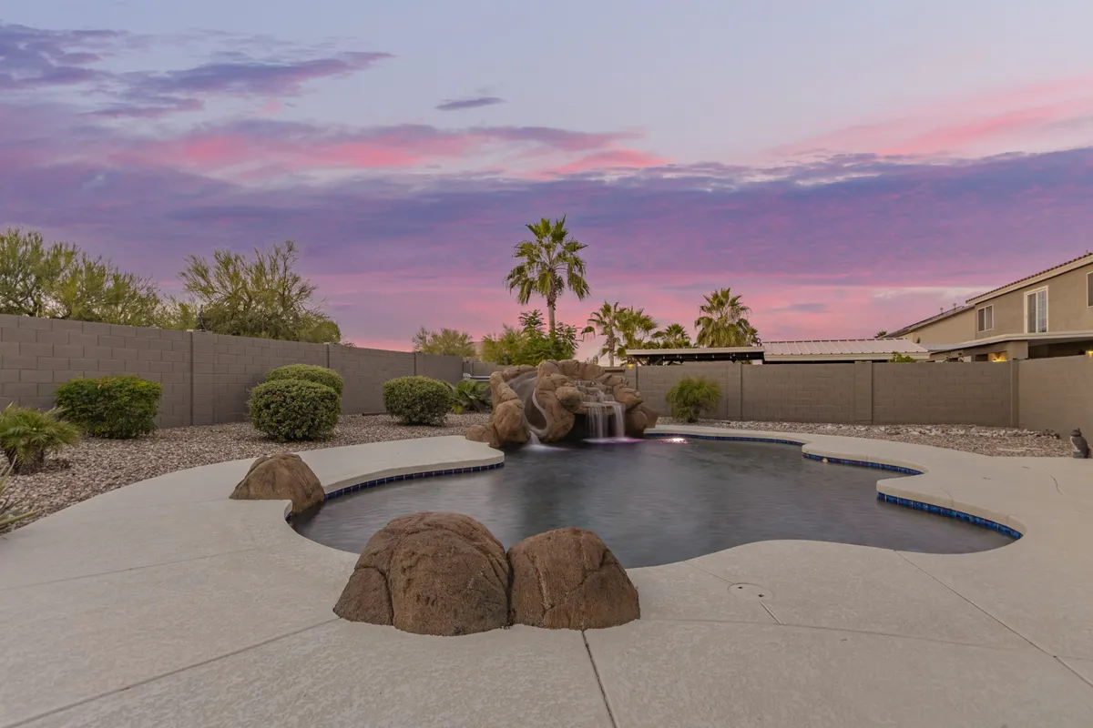 A serene backyard with a swimming pool accented by a rock waterfall and palm trees, all illuminated by the colors of a sunset sky. The inviting water hints at play, while the seating area and landscaping create a welcoming atmosphere for both adults and children.