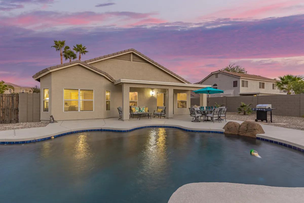 A spacious backyard scene featuring a swimming pool, comfortable patio seating, an outdoor grill, and vibrant greenery. The sunset sky provides a beautiful backdrop, suggesting the perfect environment for barbecues, social gatherings, and family relaxation.