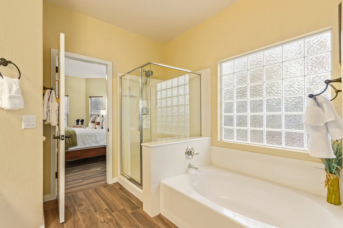 A spa-like bathroom with a luxurious soaker tub, glass-enclosed walk-in shower, and a large glass block window that brings in morning light. The interior door leads toward the bedroom, creating a private ensuite feel, while soft colors and elegant fixtures add to the calming ambiance.