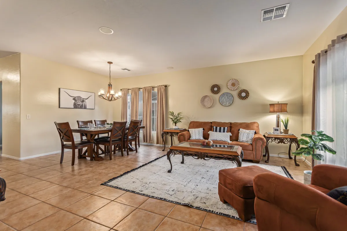 A spacious, sunlit living and dining area featuring a large table set for eight beneath a striking chandelier, with plush sofas clustered nearby for relaxation. Decorative wall plates and elegant lighting add warmth and character, while the open layout encourages seamless movement between social and dining spaces.