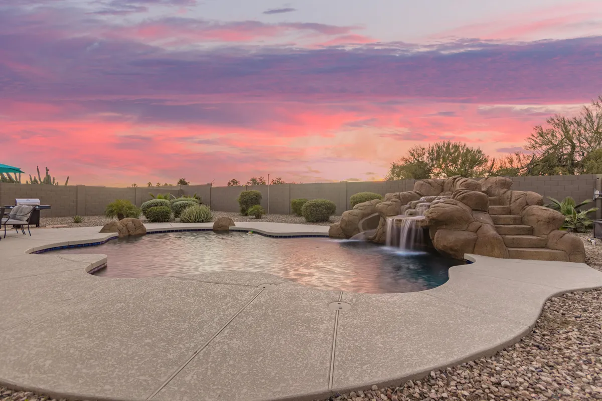 This image showcases a luxurious backyard pool complete with a waterfall feature. The sun is setting in the background, casting a warm glow over the landscaped rocks and lush plants. The space is clearly designed for both relaxation and entertainment, with areas suitable for swimming, lounging, and outdoor dining.