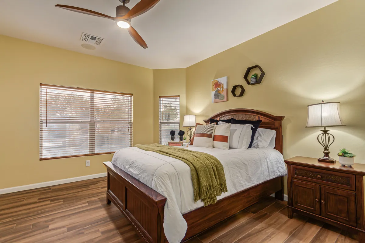 A bright, inviting bedroom featuring a queen bed with crisp linens, matching nightstands with lamps, a large window that bathes the space in natural light, and modern decorative art above the headboard. The room feels warm and restful, perfect for relaxation and privacy.