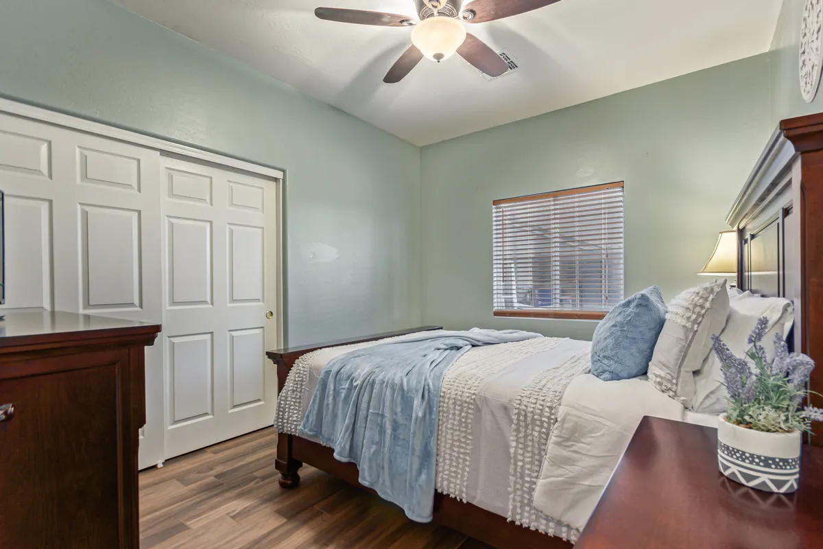 A secondary queen bedroom with a neatly made bed, soft window light streaming in, a dresser, and a decorative plant in the corner. The tranquil mood and simple, stylish furnishings create a peaceful environment suitable for guests or family members.